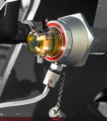 Luneta Condition Monitoring Pod fitted to a machine, with a clear oil-filled sight glass and red CMP ring; a knurled sampling probe and white tube are connected to the valve underneath, and the valve cap hangs on a short chain.